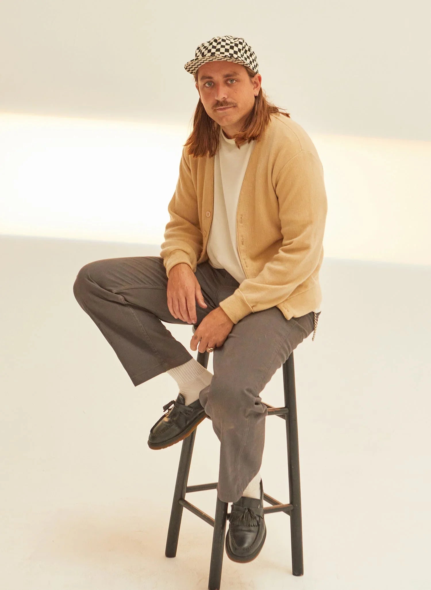 Person in beige cardigan, checkerboard cap, and loafers sitting on stool in minimalist studio