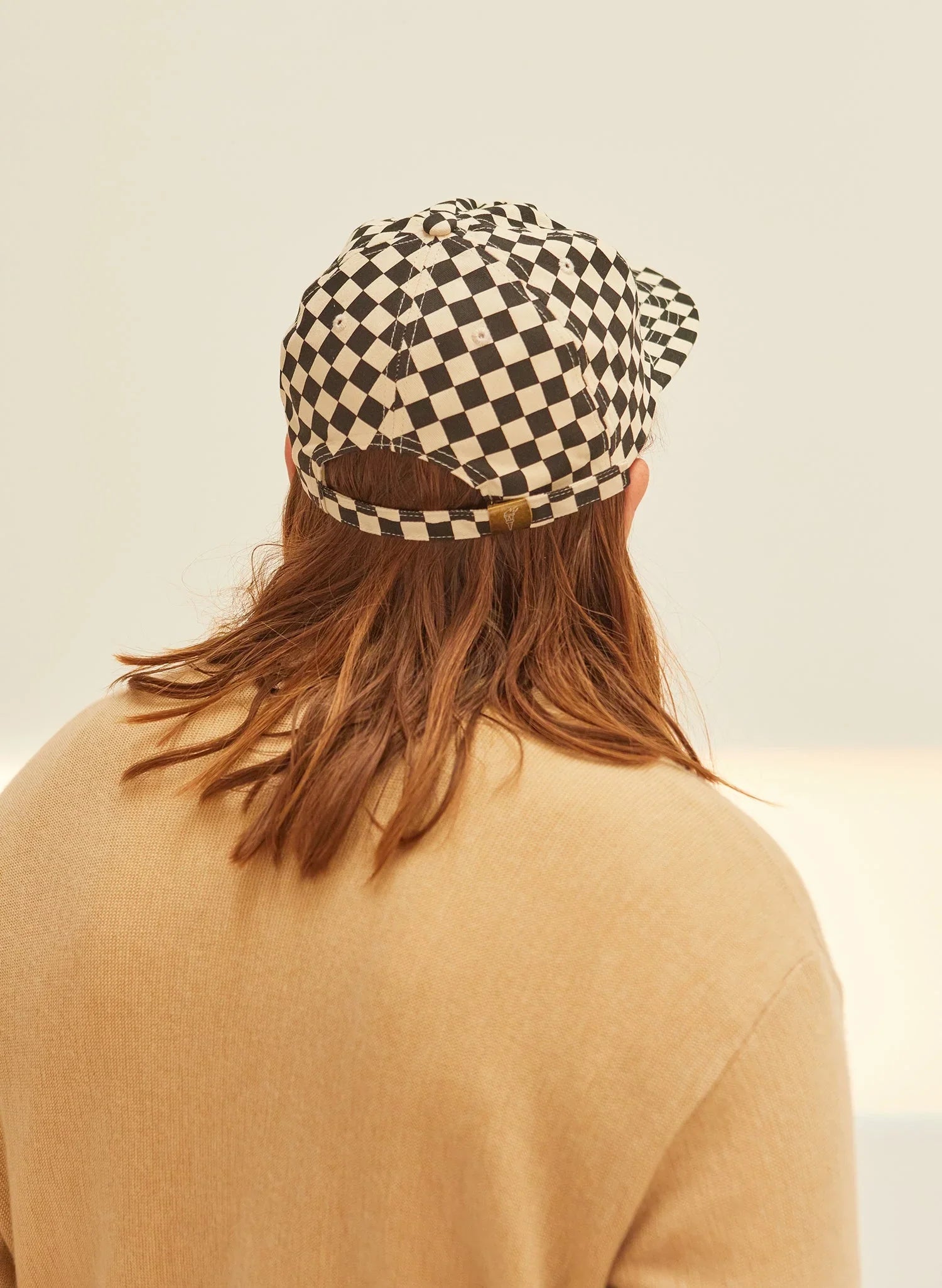 Person wearing a black and white checkerboard cap, beige top, neutral background, Qu!rk Boutique