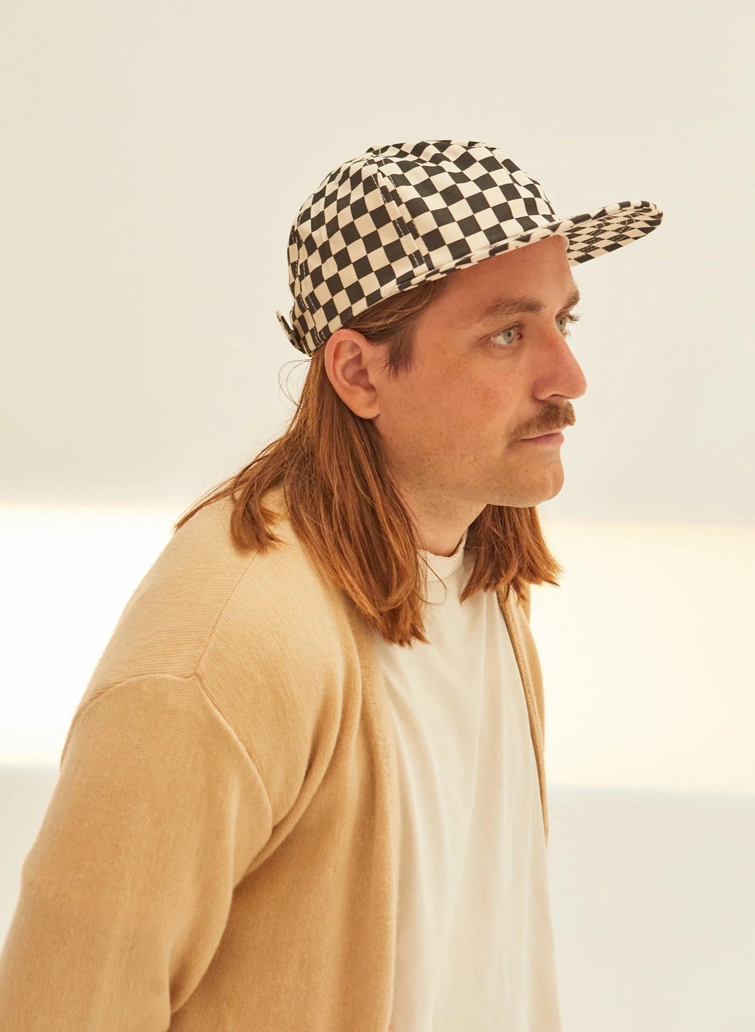 Man in neutral sweater and white shirt wearing a black-and-white checkerboard cap, minimalist background