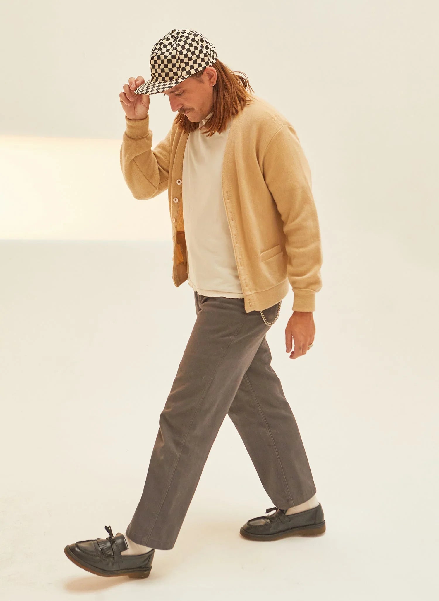 Person wearing checkerboard cap, beige cardigan, gray pants, black loafers on neutral background