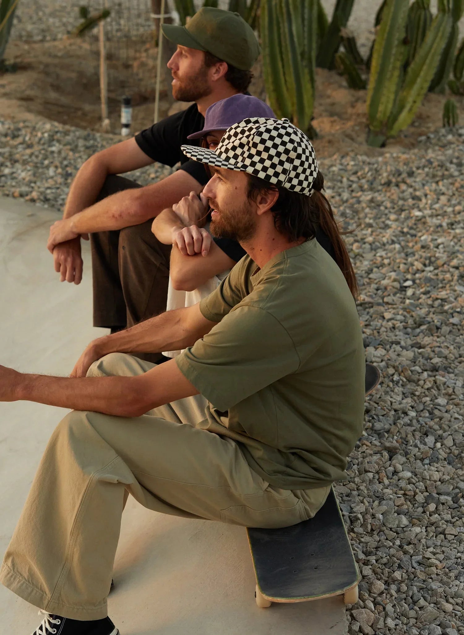 Three people wearing casual outfits and caps, including a checkerboard hat, sit on a skateboard near cacti and gravel, bathed in warm sunlight.