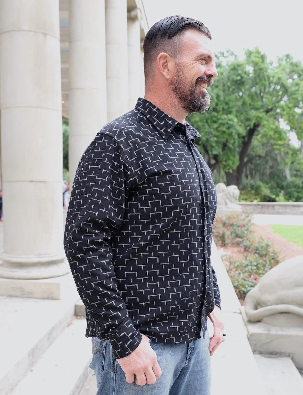 Man wearing a black geometric button-down shirt from Qu!rk Boutique, standing outdoors by columns.