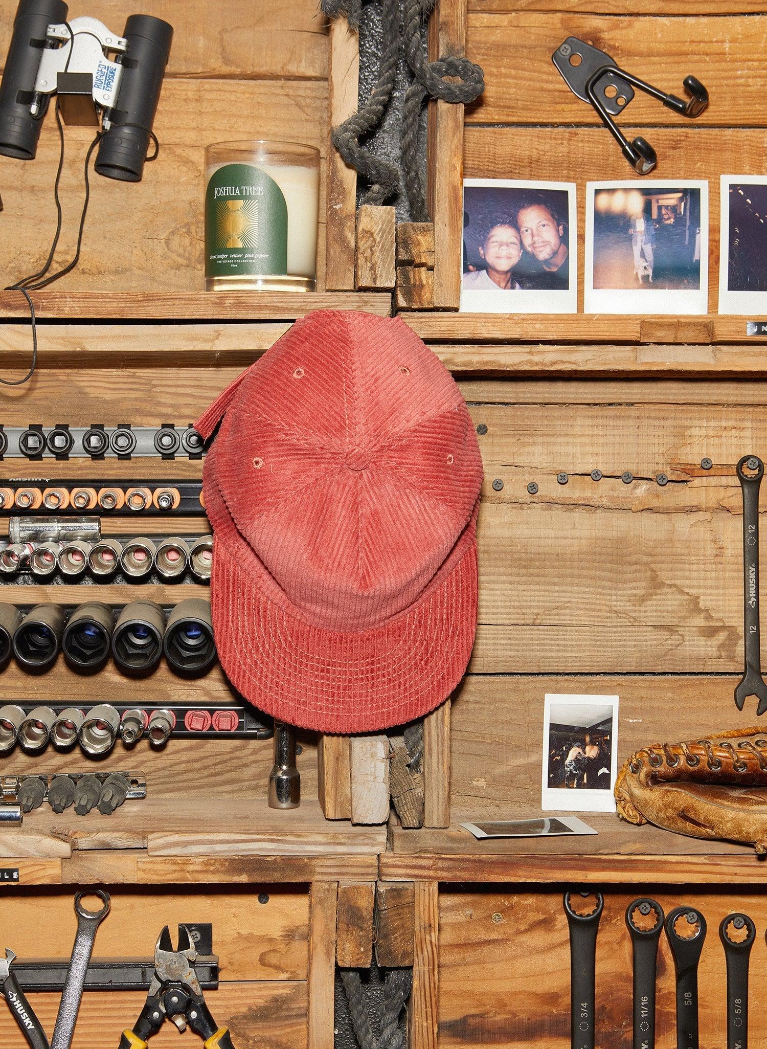 Red corduroy cap on rustic wood wall with tools, candle, and vintage photos at Qu!rk Boutique