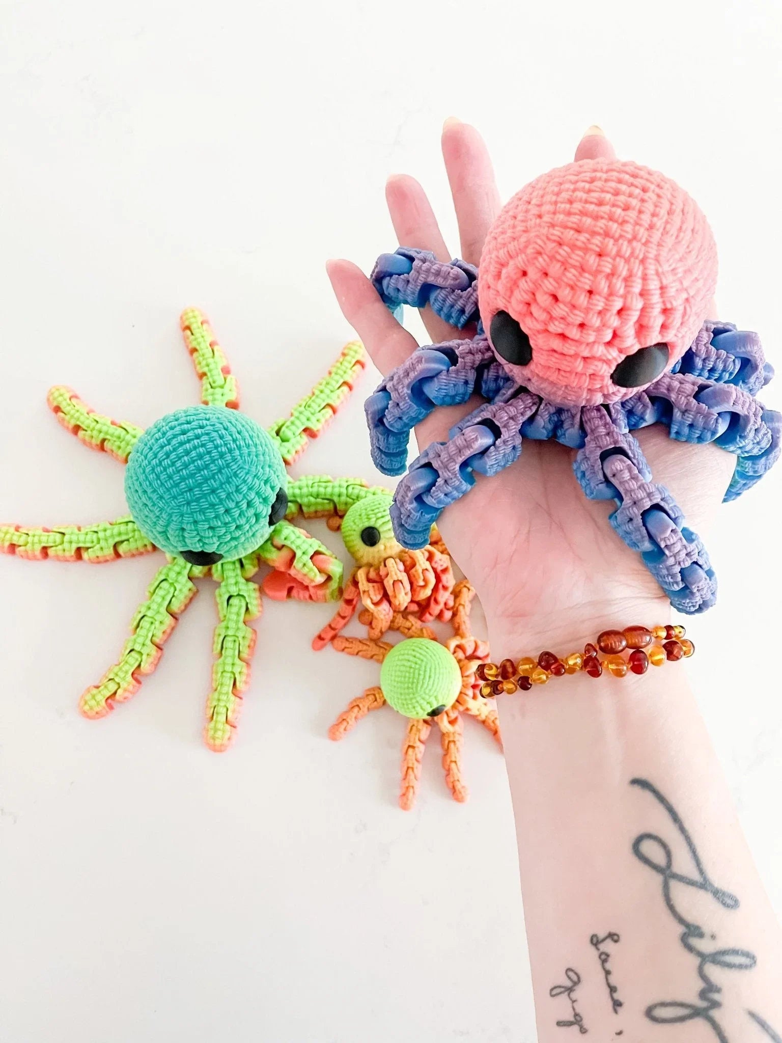 Hand holding a pink and purple crocheted octopus plush, with more colorful crocheted octopuses on white background