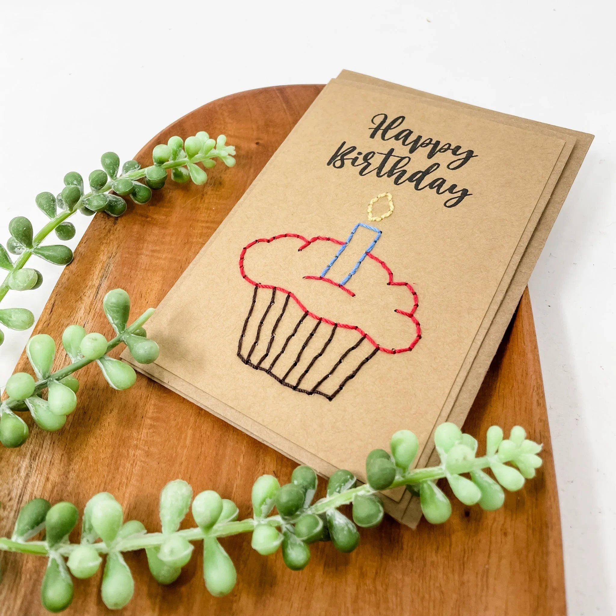 Handmade kraft birthday card with stitched cupcake and candle, styled on wood with greenery