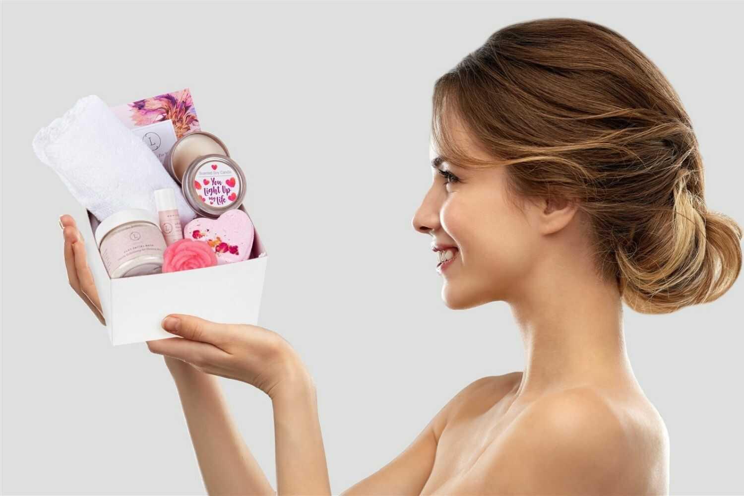 Smiling woman holding a white gift box with natural skincare products, candle, and towel
