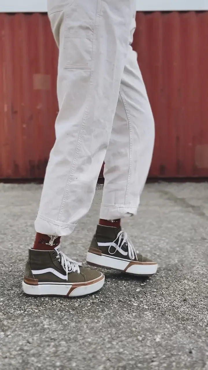 Person wearing rolled-up white pants and olive green high-top sneakers on asphalt with red wall background