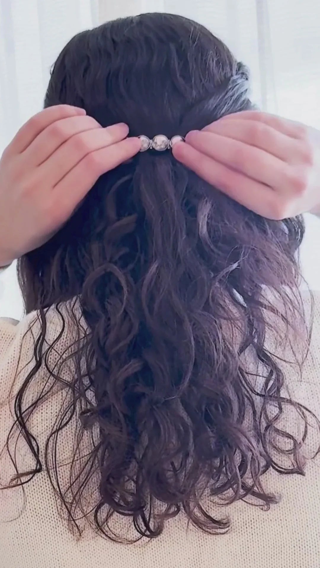 Person with long curly dark hair securing silver hair clip on half-up hairstyle indoors