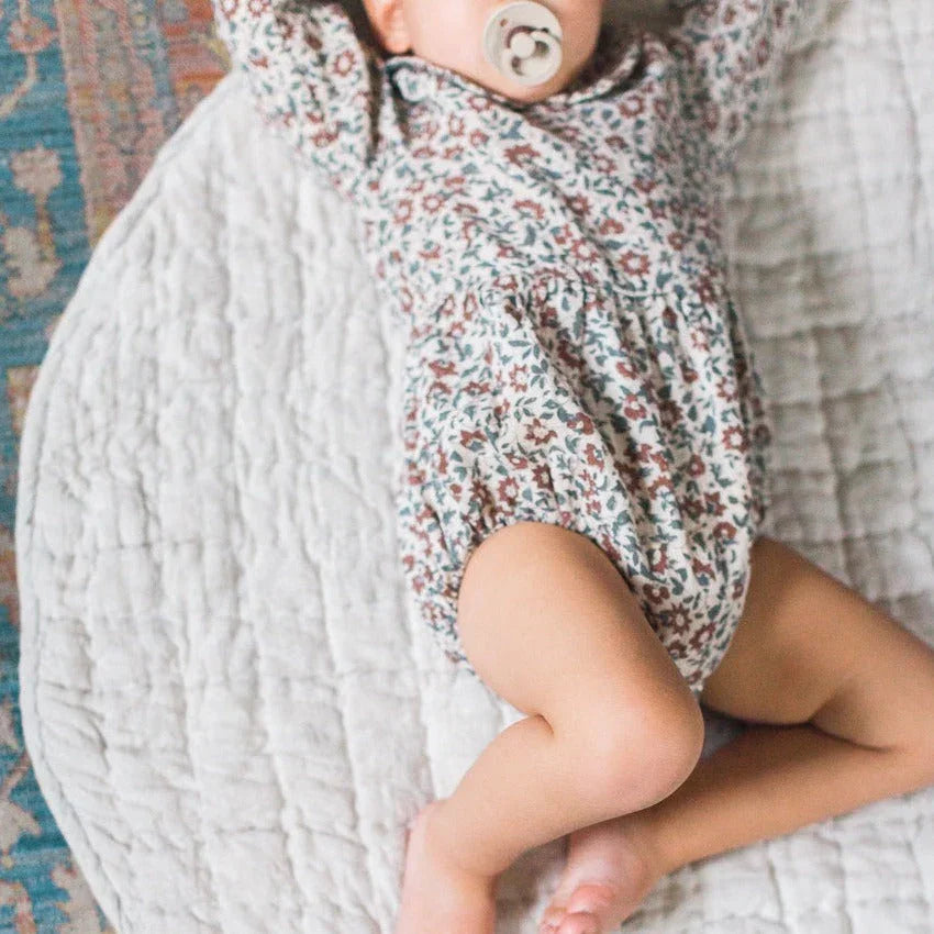 Baby in floral romper with pacifier lying on soft quilt, Qu!rk Boutique sustainable style