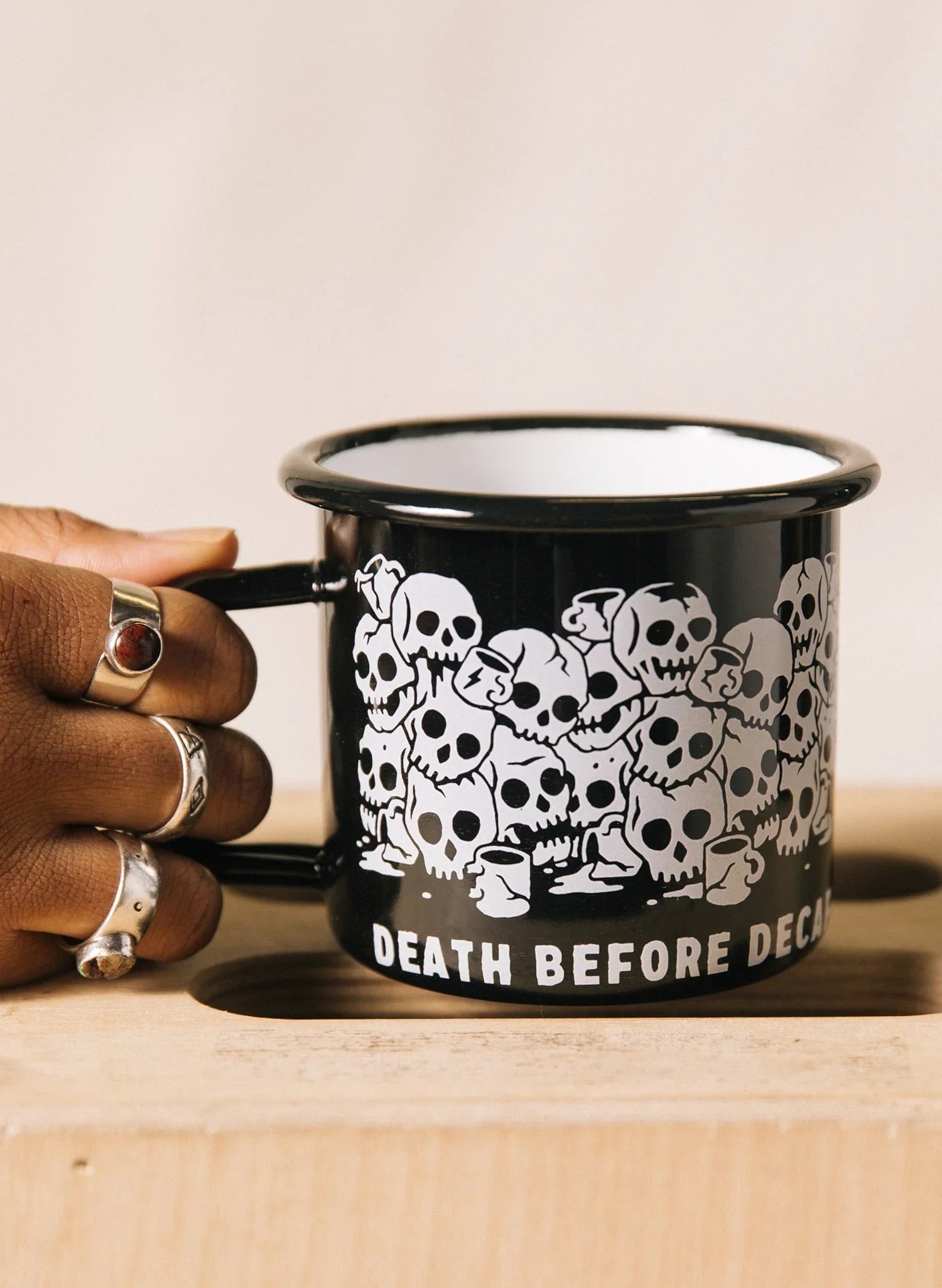 Black enamel mug with white skull illustrations and 'Death Before Decaf' text, held by hand