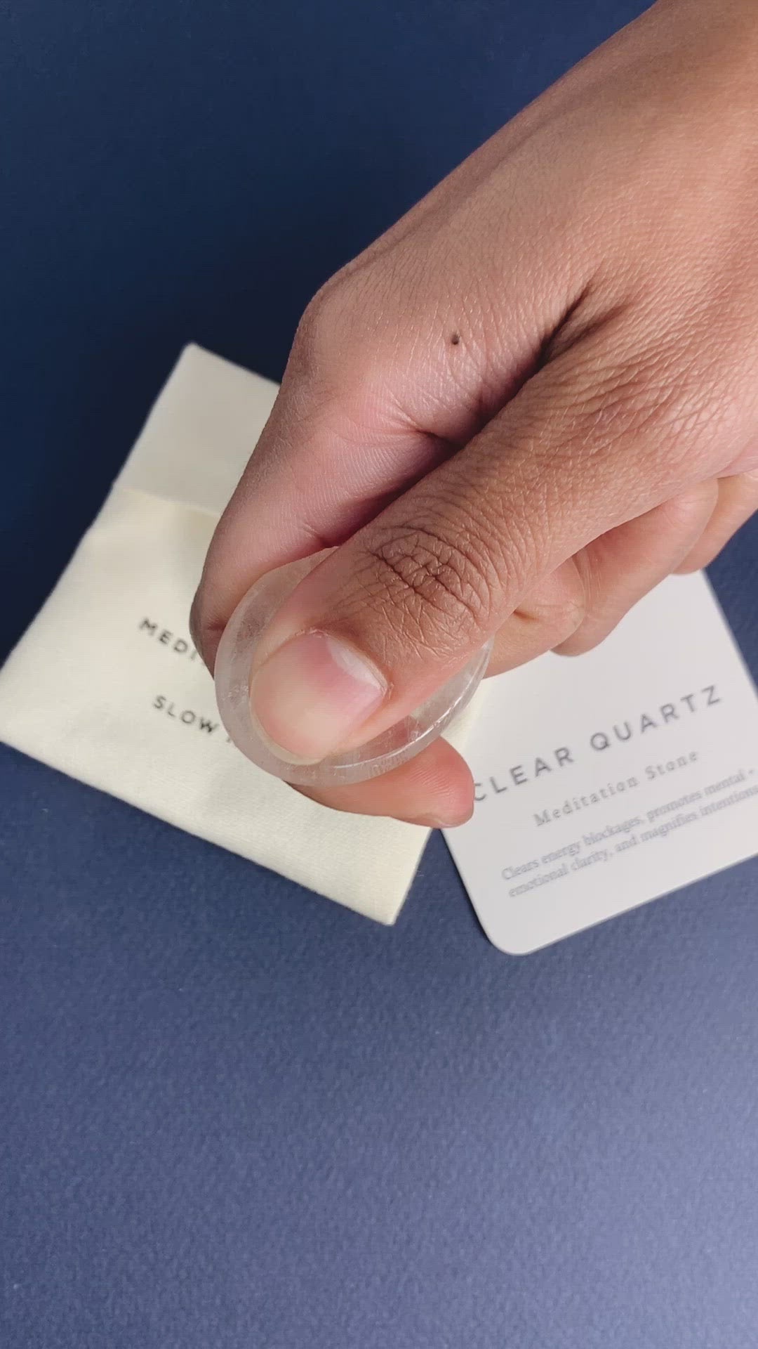 Hand holding a clear quartz meditation stone over cloth pouch and clear quartz information card on blue surface