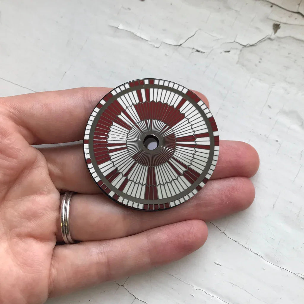 Hand holding a circular enamel pin with red and white radial geometric pattern and central hole