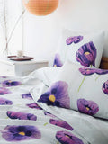 White bedding set featuring purple poppy flower prints on bed with white nightstand and candle