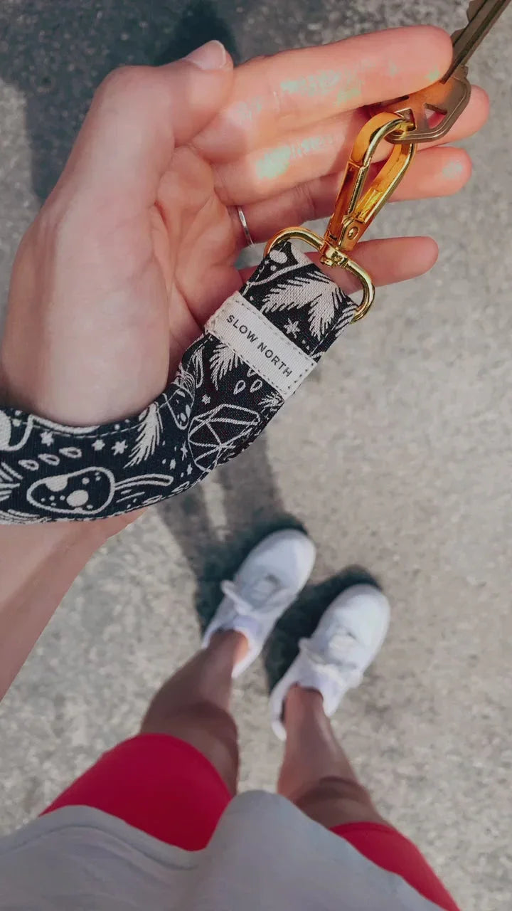 Hand holding black patterned Slow North keychain with gold clasp, person wearing red shorts and white sneakers