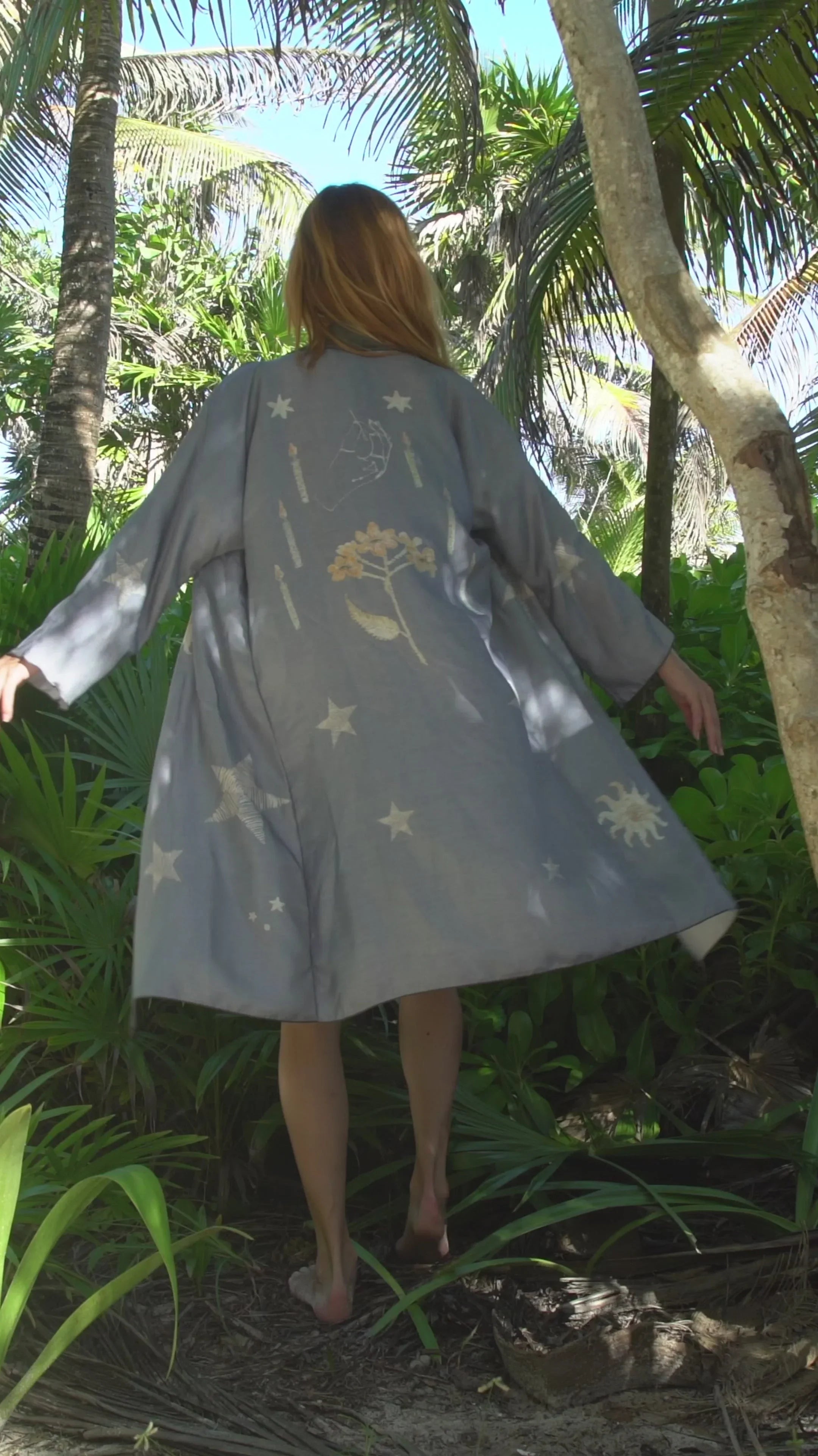 Woman wearing a loose gray robe with celestial and botanical prints, walking barefoot in a tropical forest