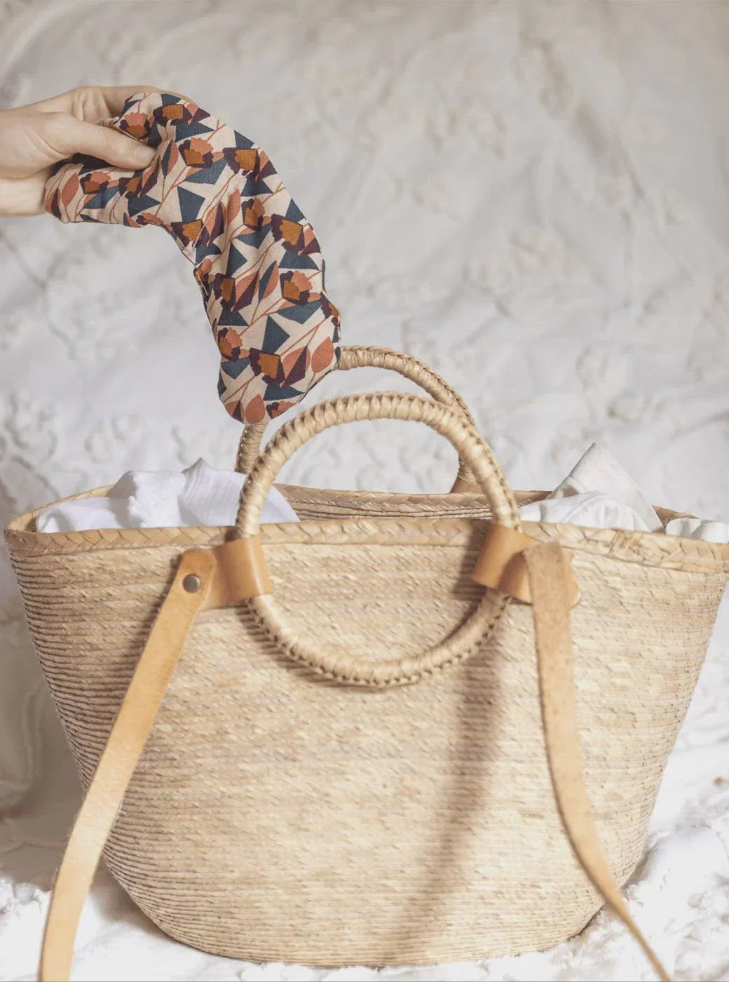 Hand holding floral sleep mask above woven straw tote bag with leather straps on white fabric
