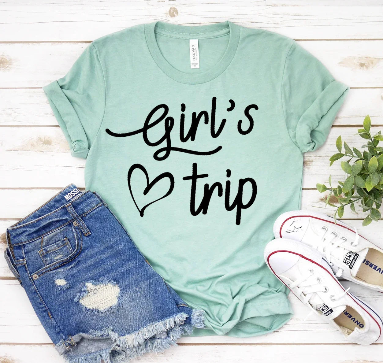 Mint green Girl's Trip graphic t-shirt with rolled sleeves, distressed denim shorts, and white sneakers on white wooden background
