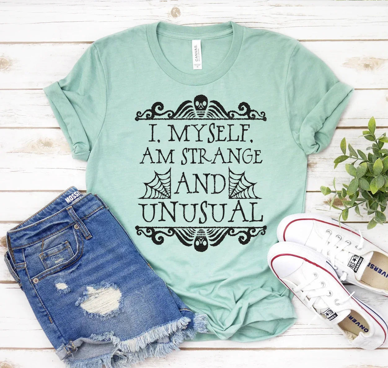 Light green t-shirt with black skull and spider web design and text I, myself, am strange and unusual, paired with ripped denim shorts and white sneakers