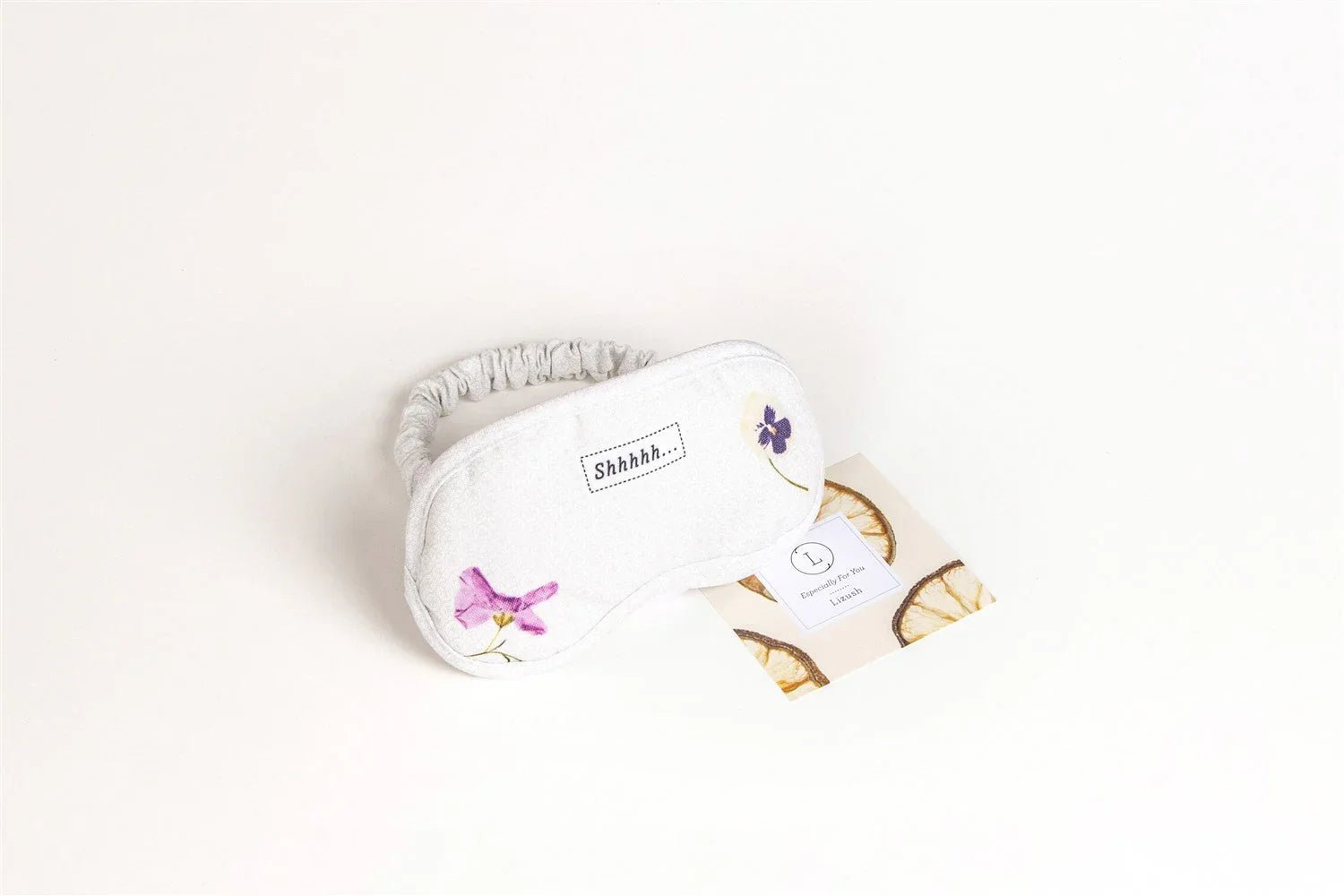 White sleep mask with elastic strap, floral prints, and 'Shhhhh...' text on a light background