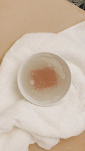 White bowl with brown soap soaking in water on a white towel and wooden surface
