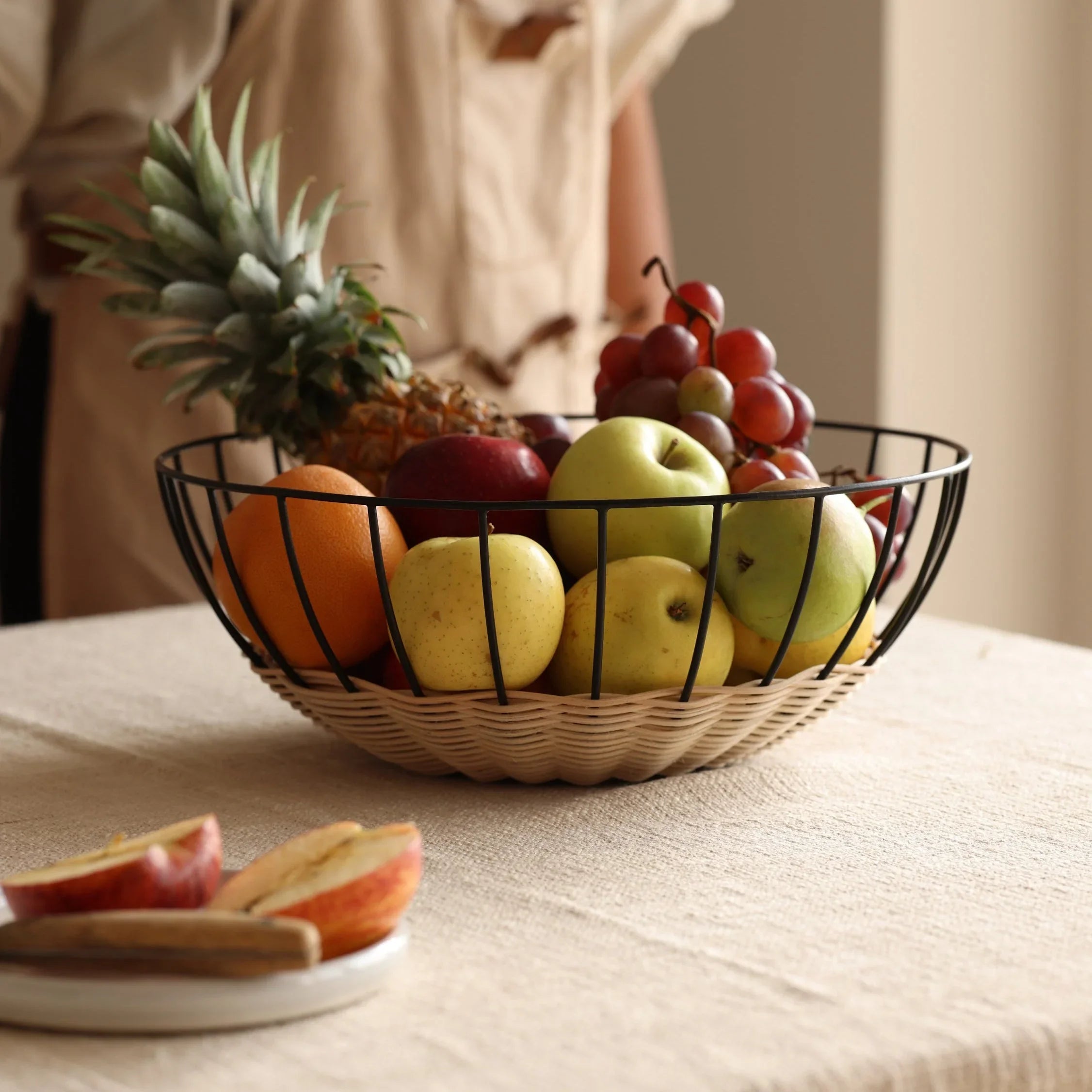 Modern wire fruit basket with apples, oranges, grapes, pineapple on textured tablecloth