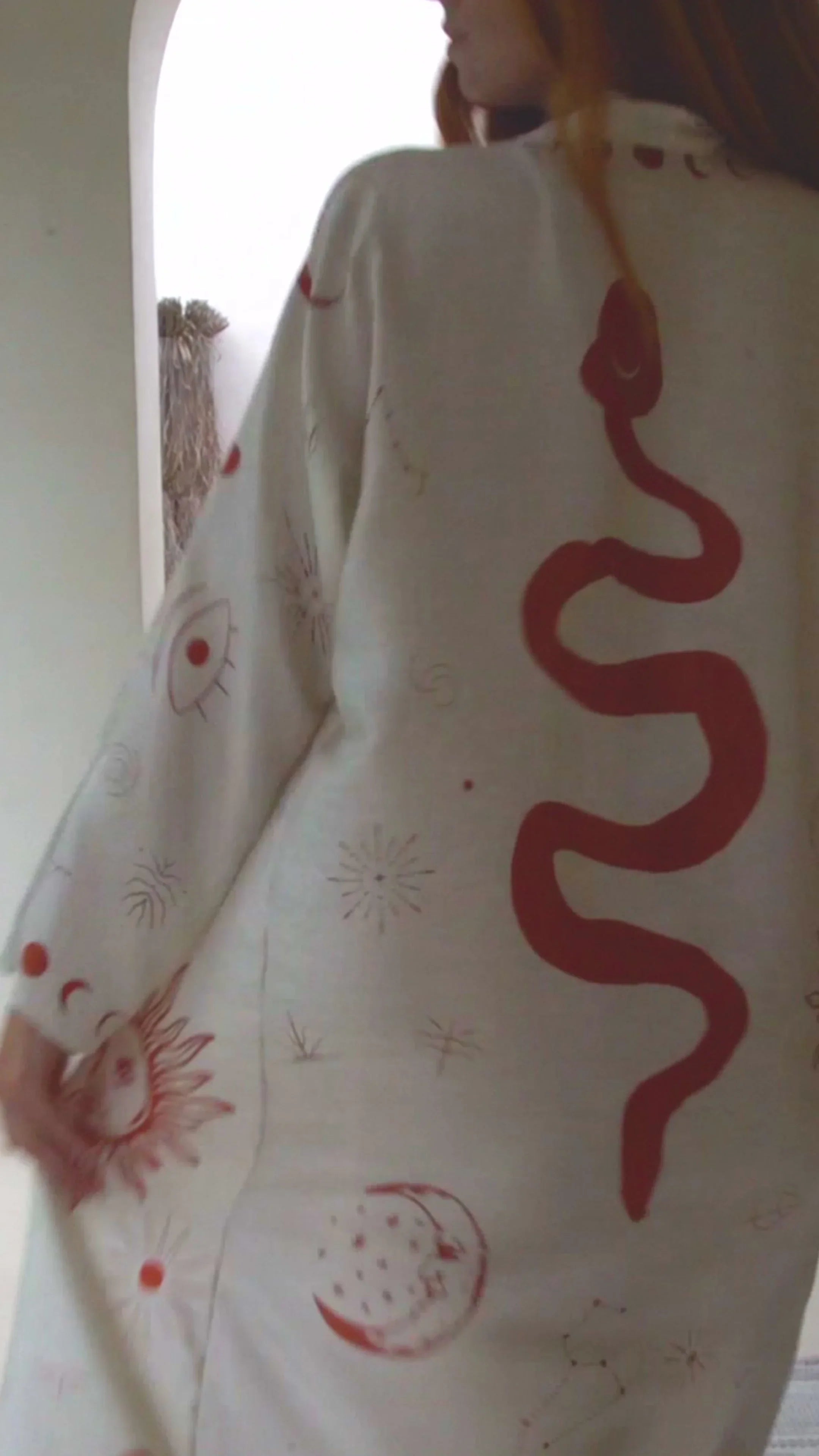 Woman wearing a white garment with red abstract designs including a snake, sun, moon, and eyes