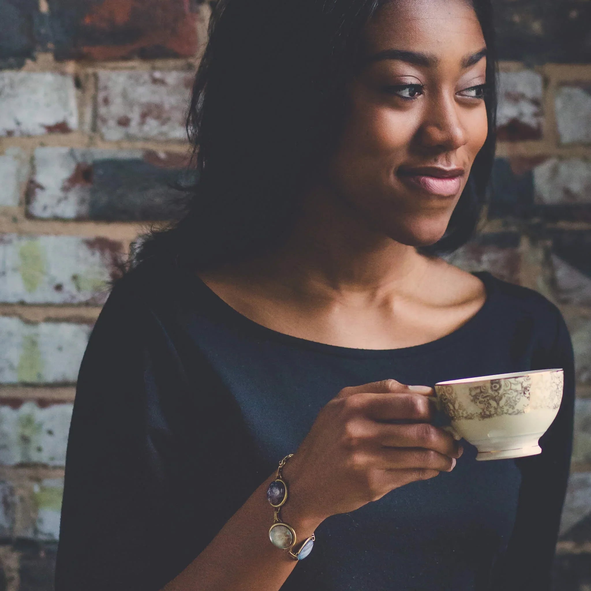 Woman in black top wearing an artisan bracelet, holding a teacup, brick wall background