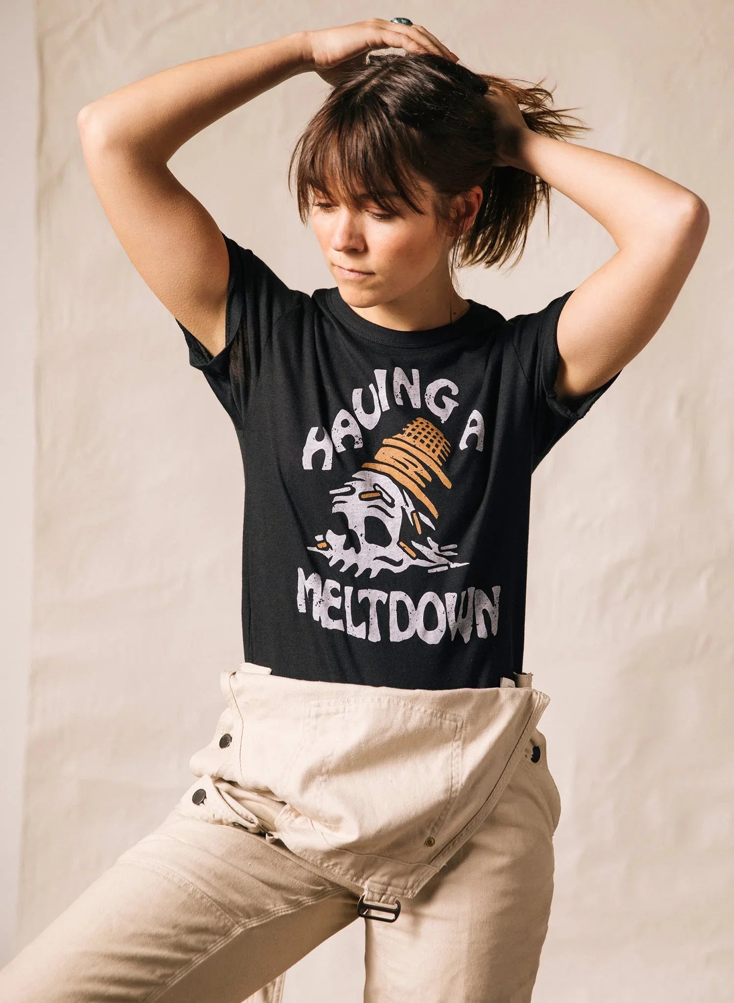 Woman wearing black graphic tee with skull and 'Having a Meltdown' text, beige pants, and hand in hair