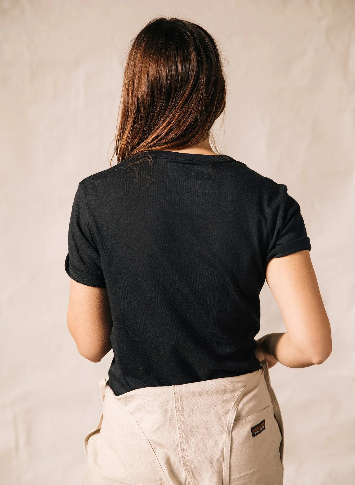 Woman with long hair wearing a black rolled sleeve tee and beige pants against neutral background
