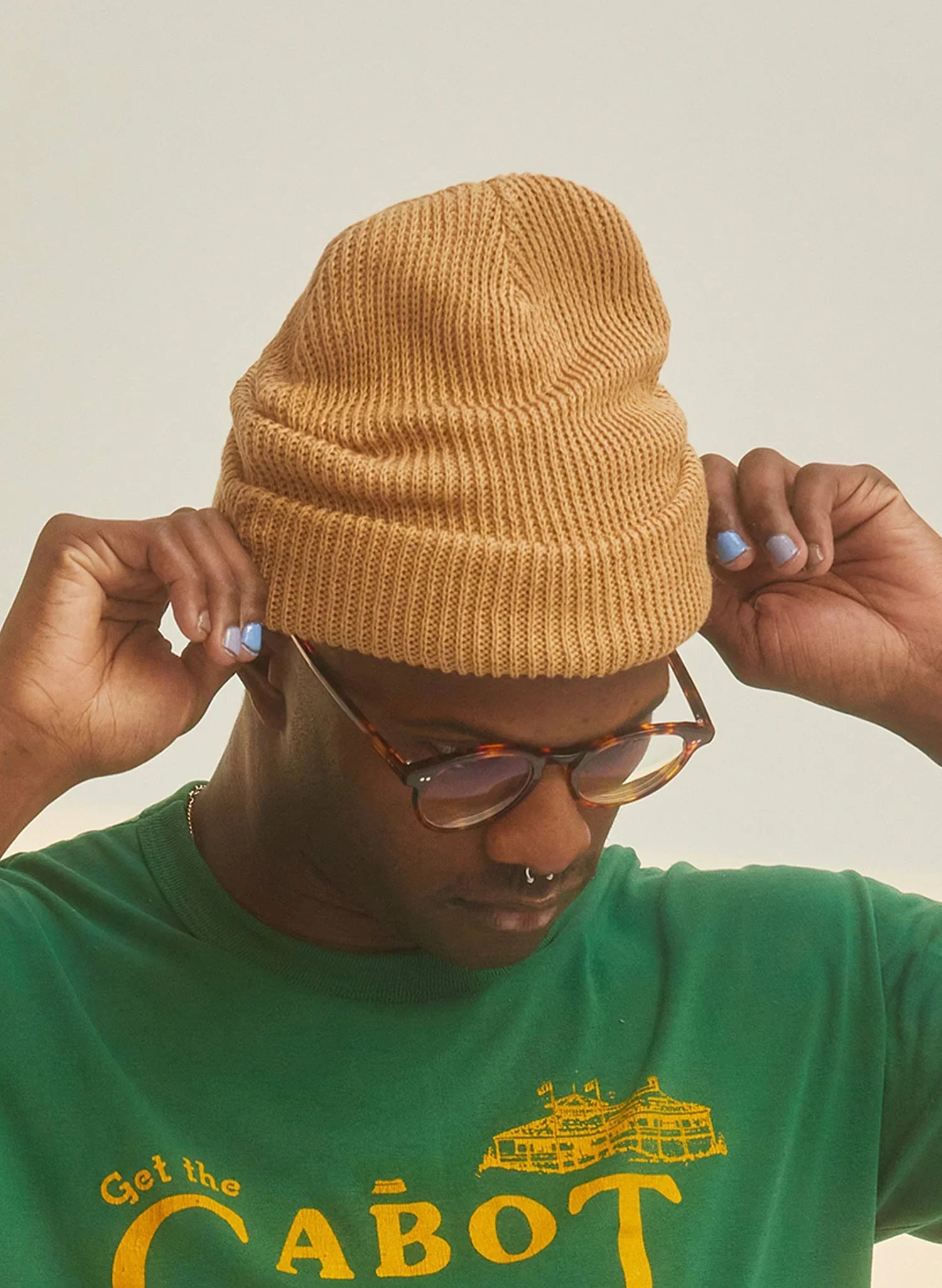 Person wearing a tan hemp beanie and green graphic tee, adjusting glasses, casual boutique style