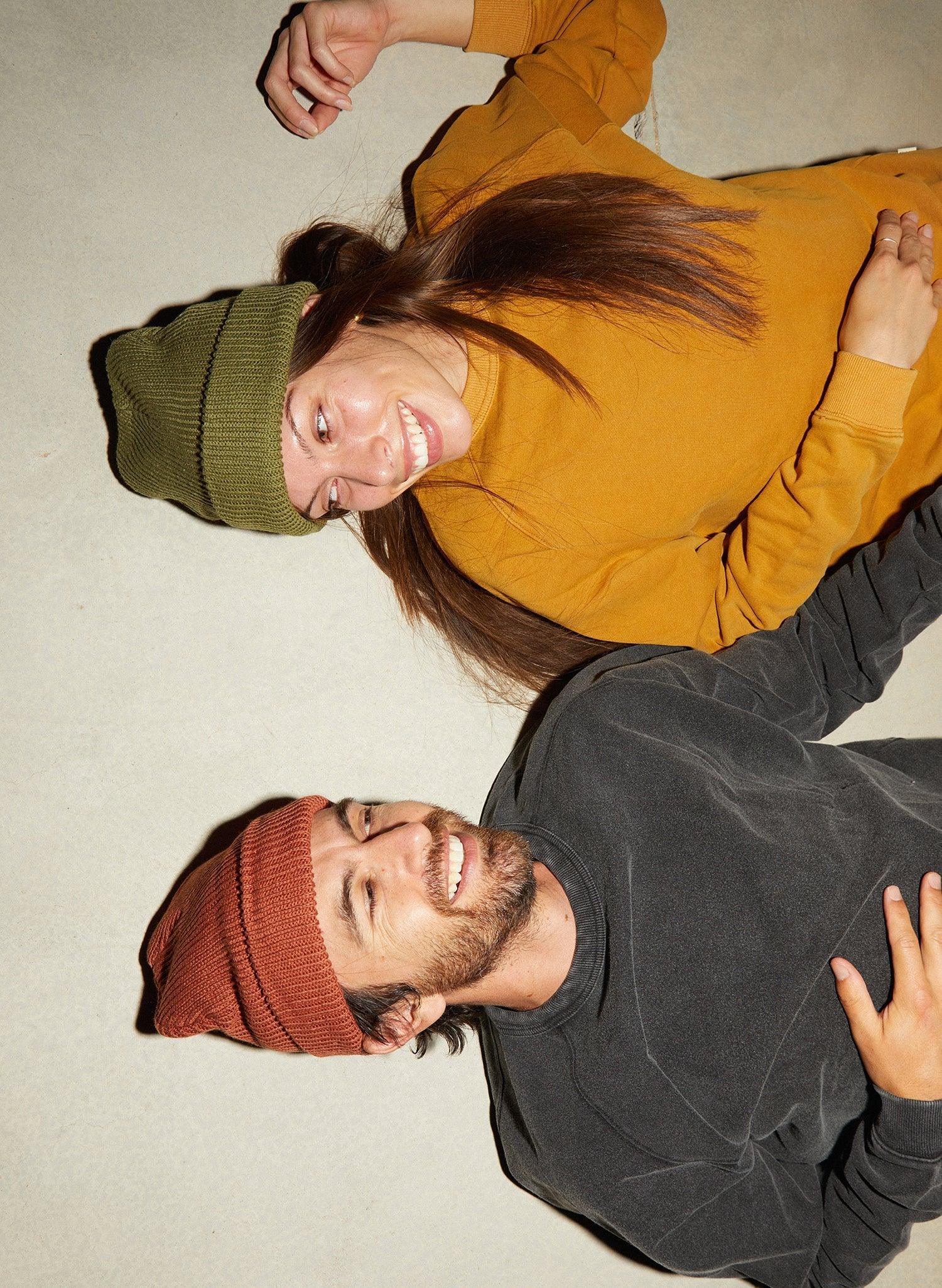 Smiling man in rust beanie and woman in green beanie, cozy sweaters, lying on light surface