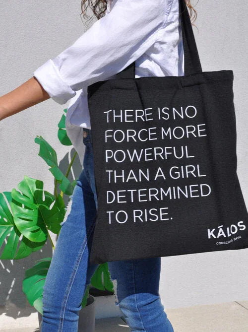 Black tote bag with empowering white text quote carried by person in white shirt and jeans against a gray wall with green plant