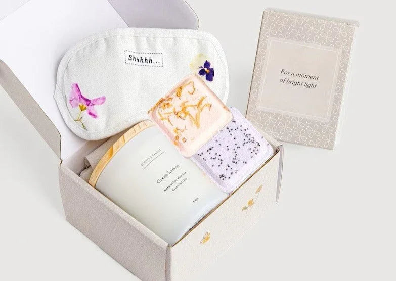 Open gift box with green lemon scented candle, floral sleep mask, two exfoliating soap bars, and card saying 'For a moment of bright light'