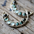 Artist-designed crescent moon earrings with green gemstone cabochons on rustic wood background