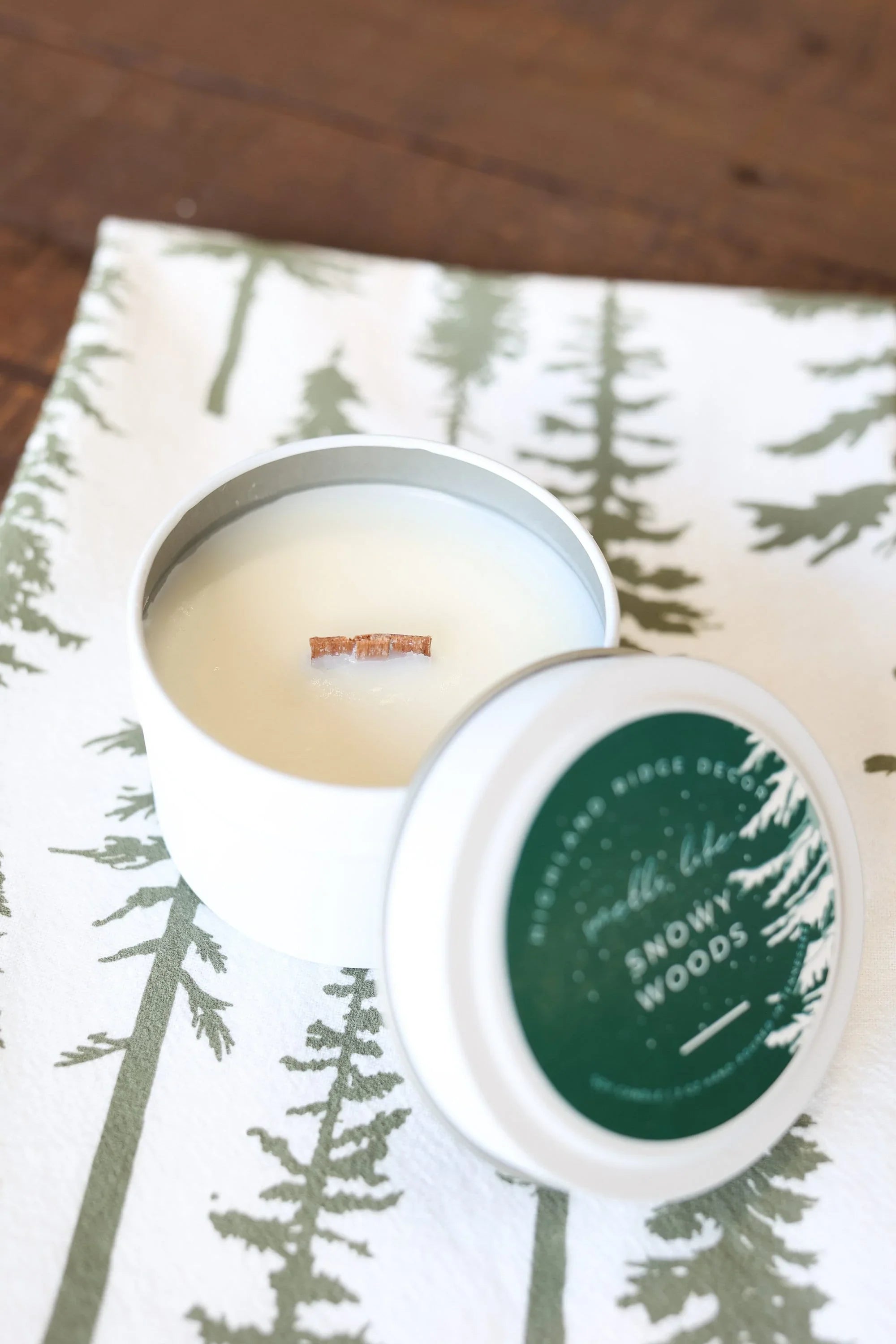 Snowy Woods soy candle in white tin with wooden wick, on pine forest print cloth, eco-friendly gift