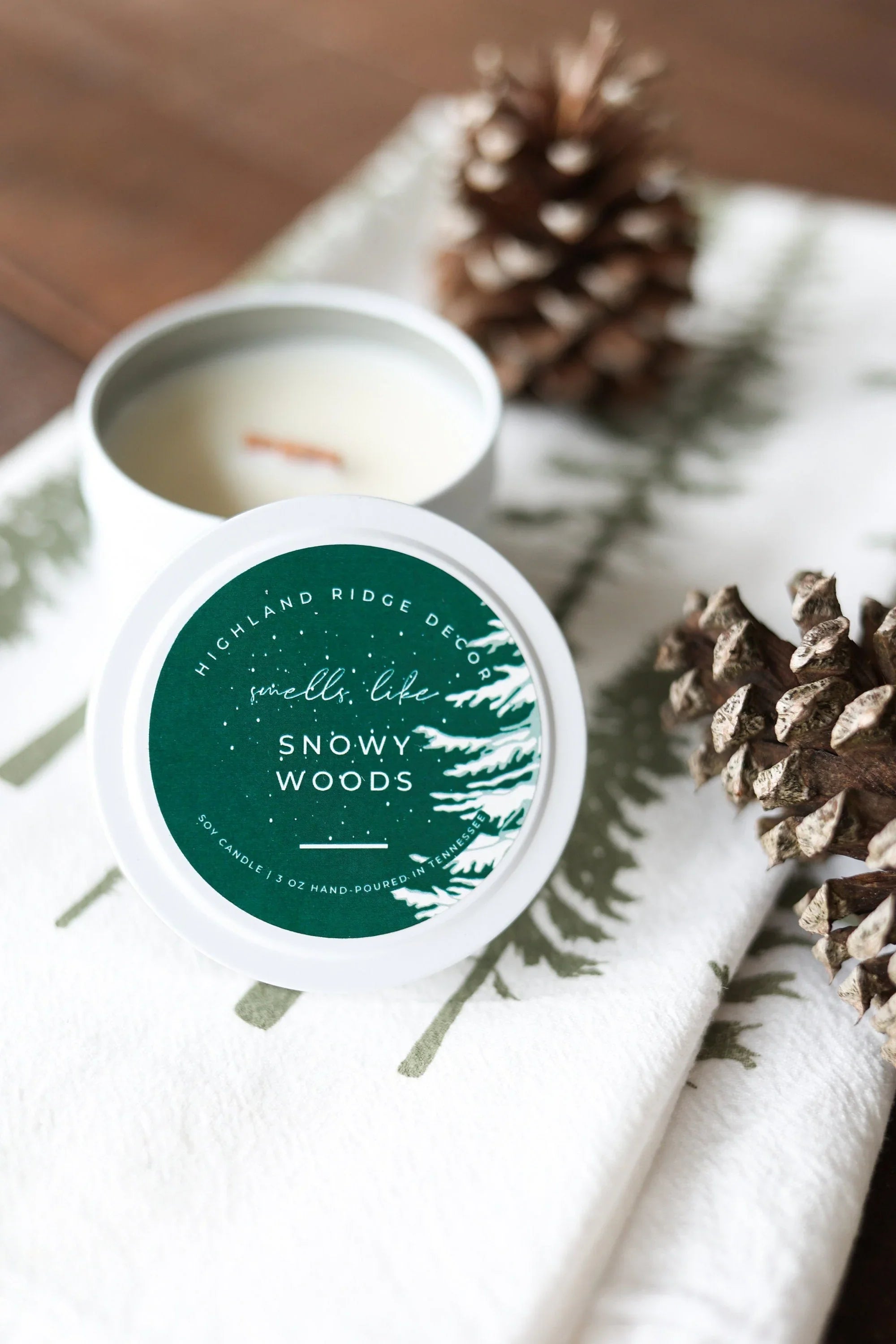 Snowy Woods soy candle tin on pine tree print cloth with pinecones, winter gift decor.