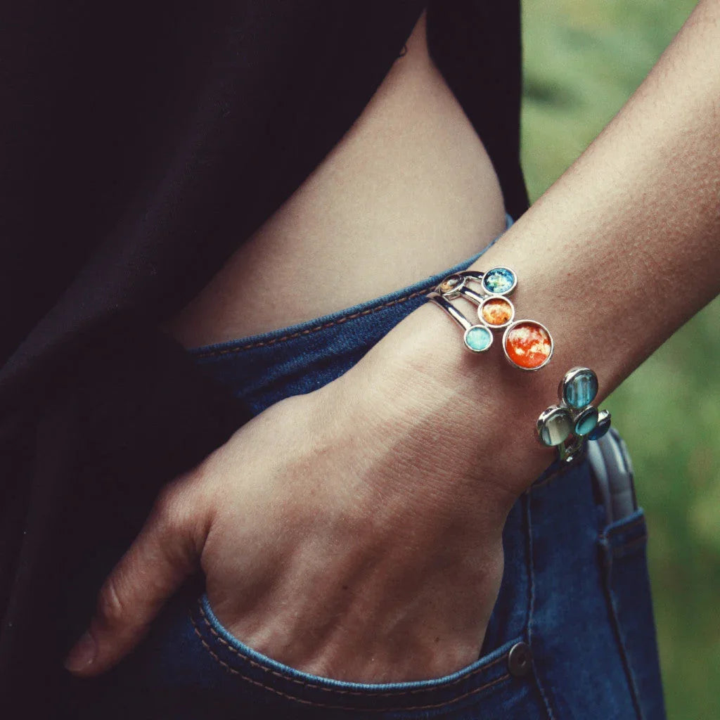 Close-up of wrist wearing a silver bracelet with colorful planet charms over blue jeans and black top