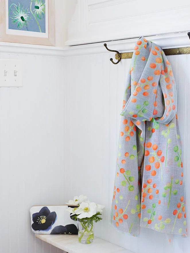 Light gray scarf with orange and green berry pattern hanging on brass hooks in white room with floral print and flowers
