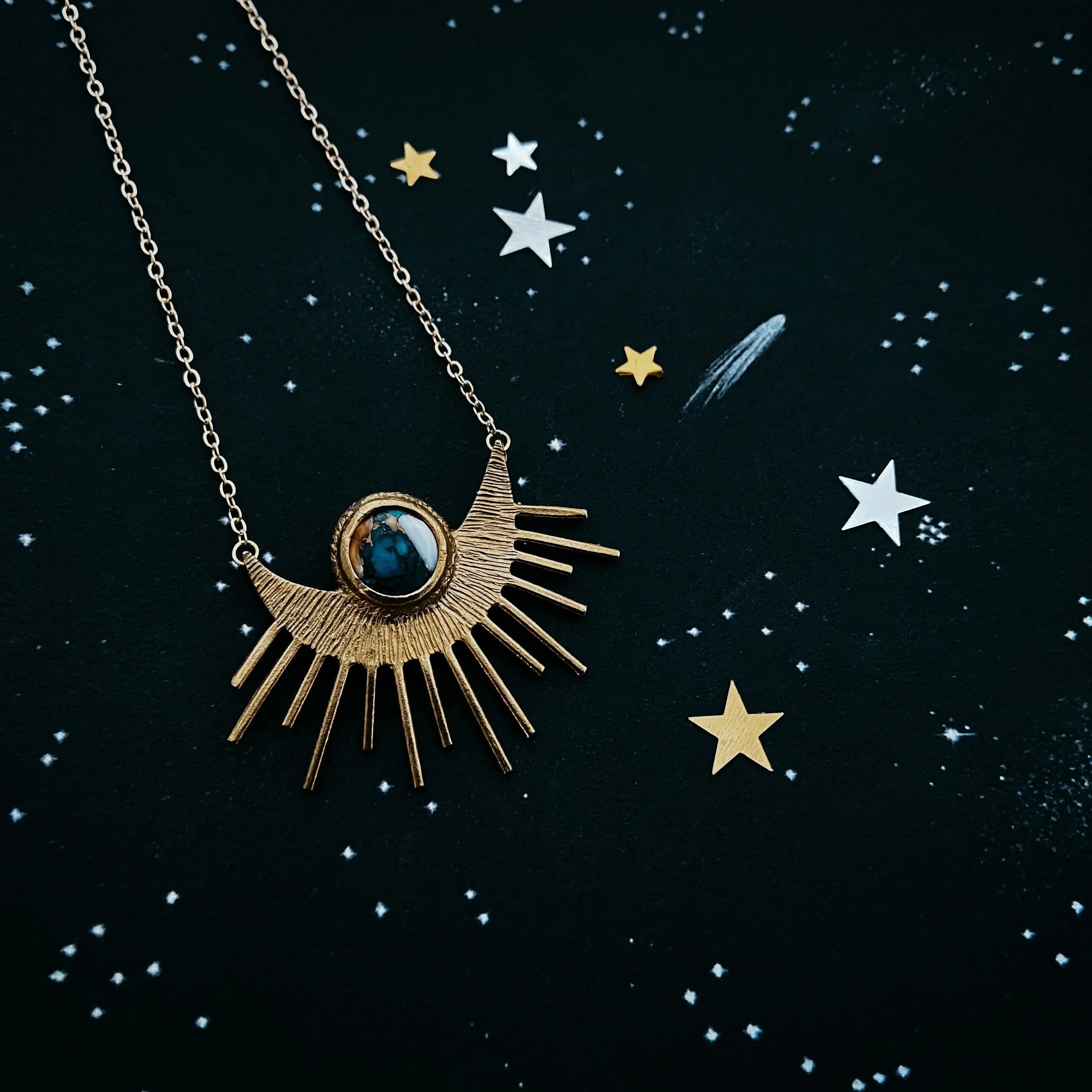 Gold sunburst necklace with blue stone pendant on black starry background with gold and silver stars