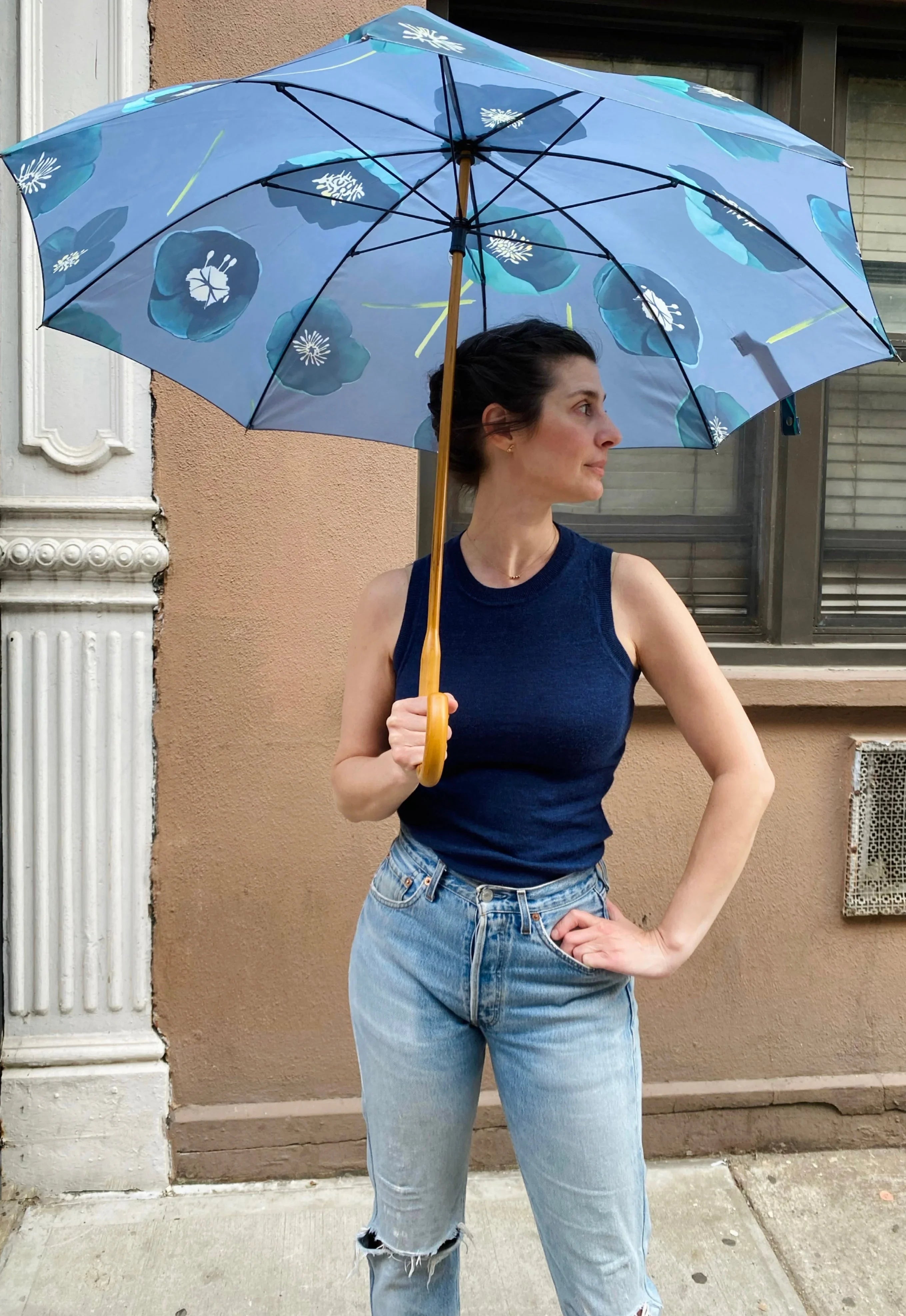 Woman in navy sleeveless top and ripped jeans holding blue floral print umbrella on city sidewalk