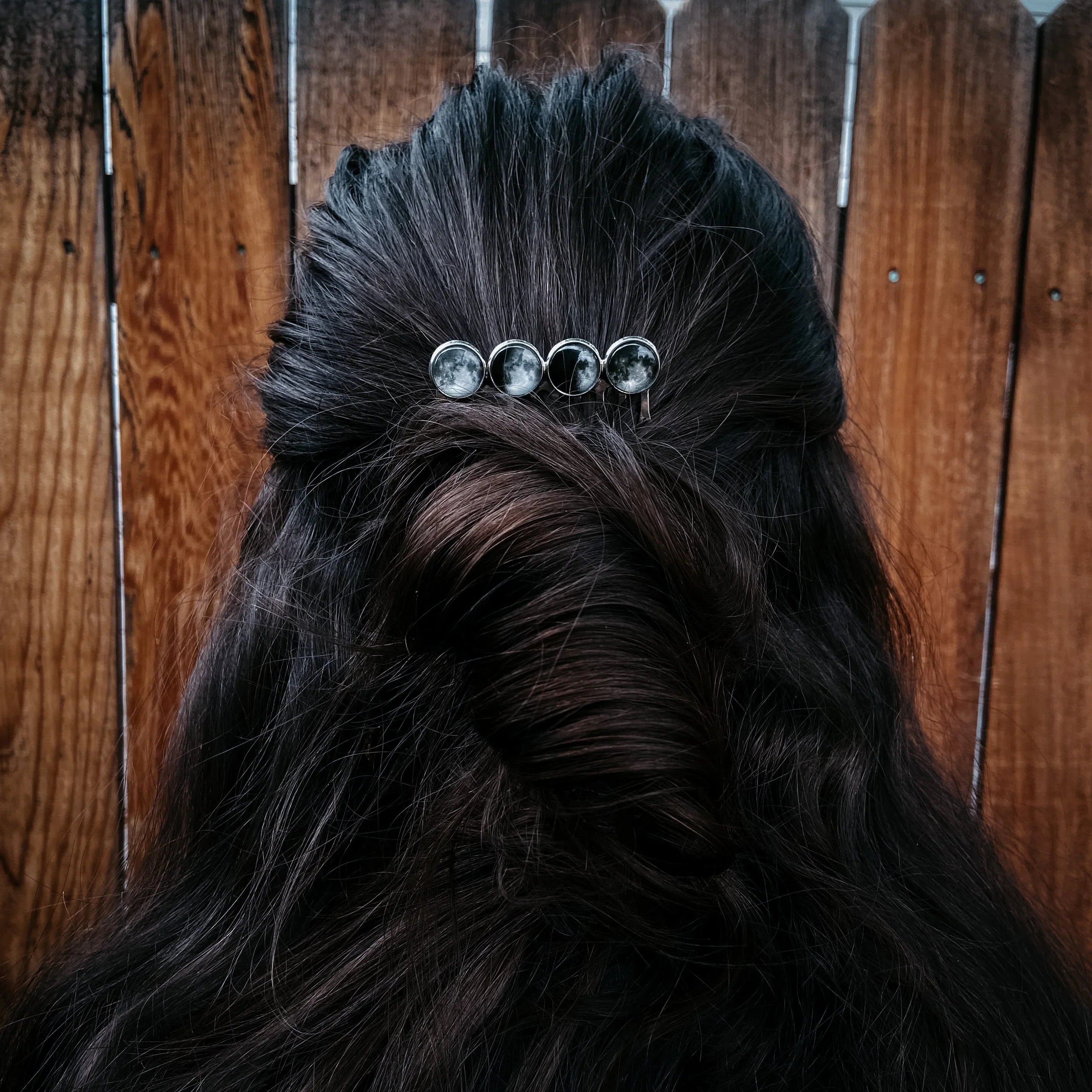 Dark brown hair styled with a twisted half-updo secured by a silver moon phase hair clip against a wooden fence background