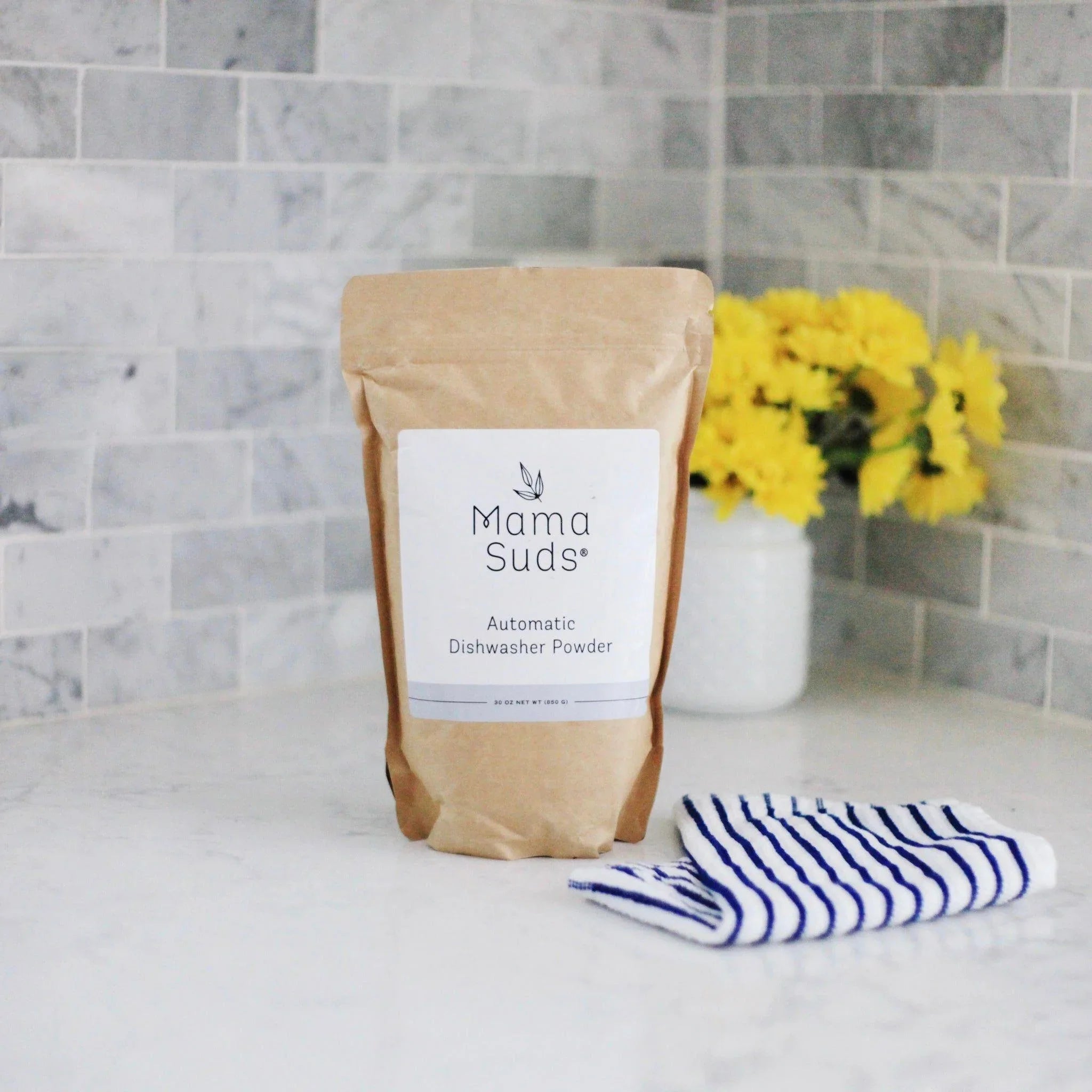 Mama Suds automatic dishwasher powder in kraft bag on kitchen counter with flowers and towel