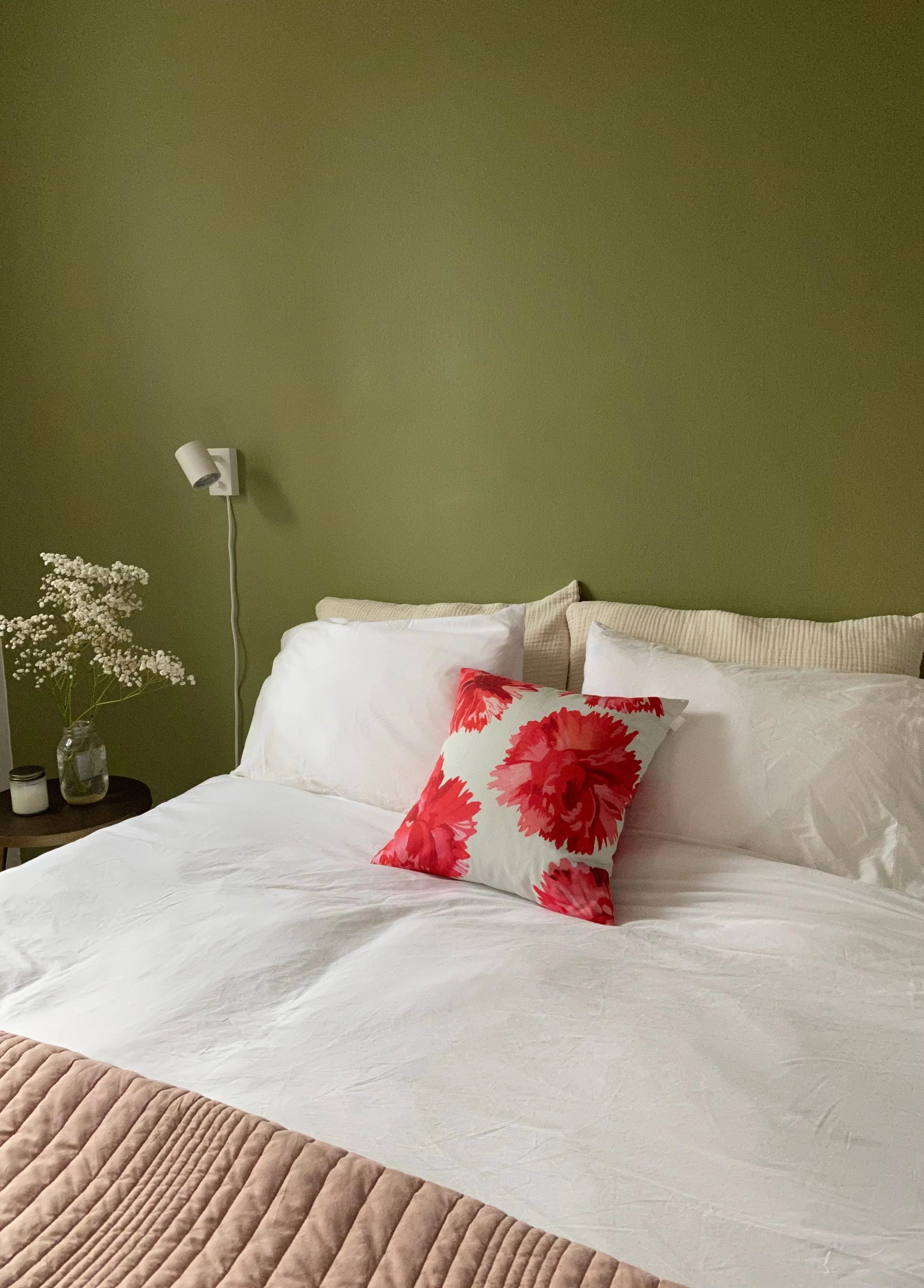 Minimalist bedroom with olive green wall, white bedding, red floral accent pillow, and bedside table with flowers