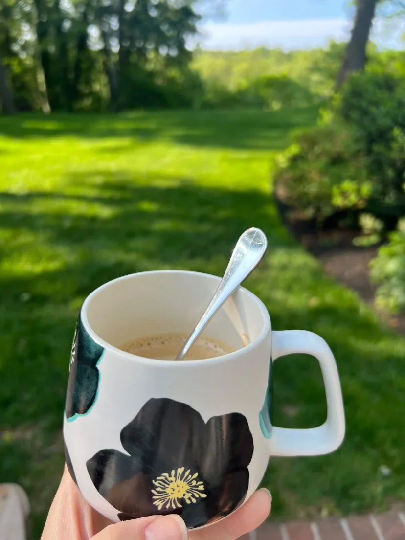 Hand holding white ceramic mug with black flower design and spoon, with green grassy backyard in sunlight