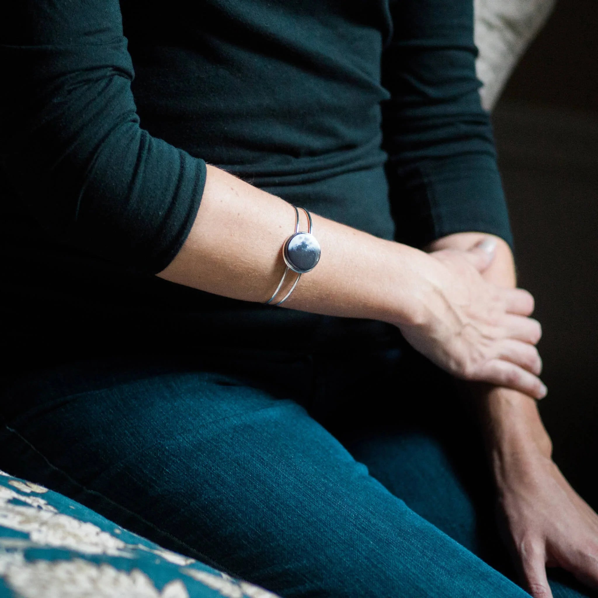 Close-up of a person wearing a silver moon phase cuff bracelet on forearm, dressed in black top and blue jeans