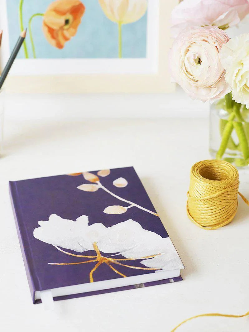 Purple journal with white flower design, yellow twine spool, and pink ranunculus flowers on white table