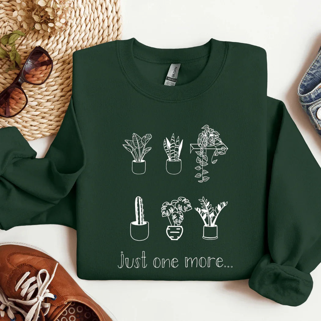 Dark green sweatshirt with white line drawings of six potted plants and the text 'just one more...'; styled with sunglasses, woven hat, brown shoes, and jeans.