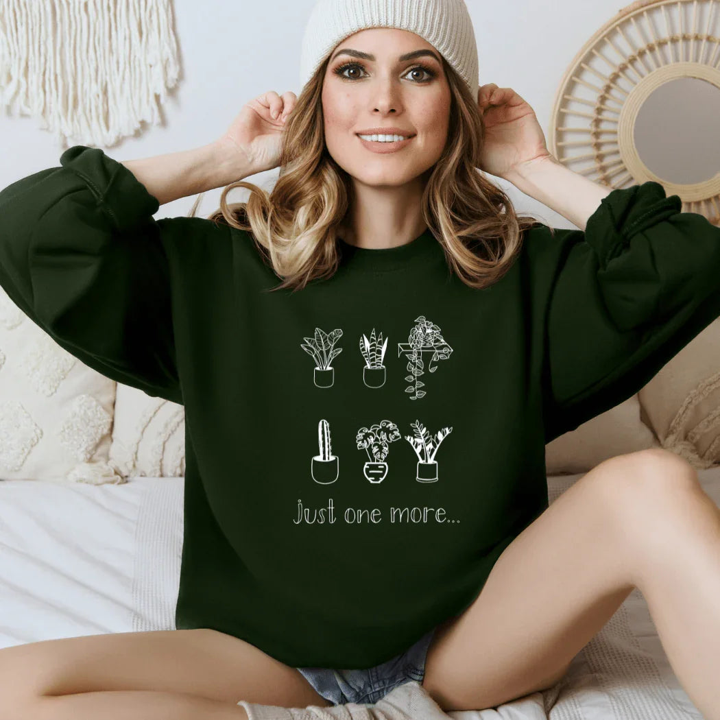 Woman in a dark green plant print sweatshirt reading 'just one more,' cozy bedroom setting, Qu!rk Boutique