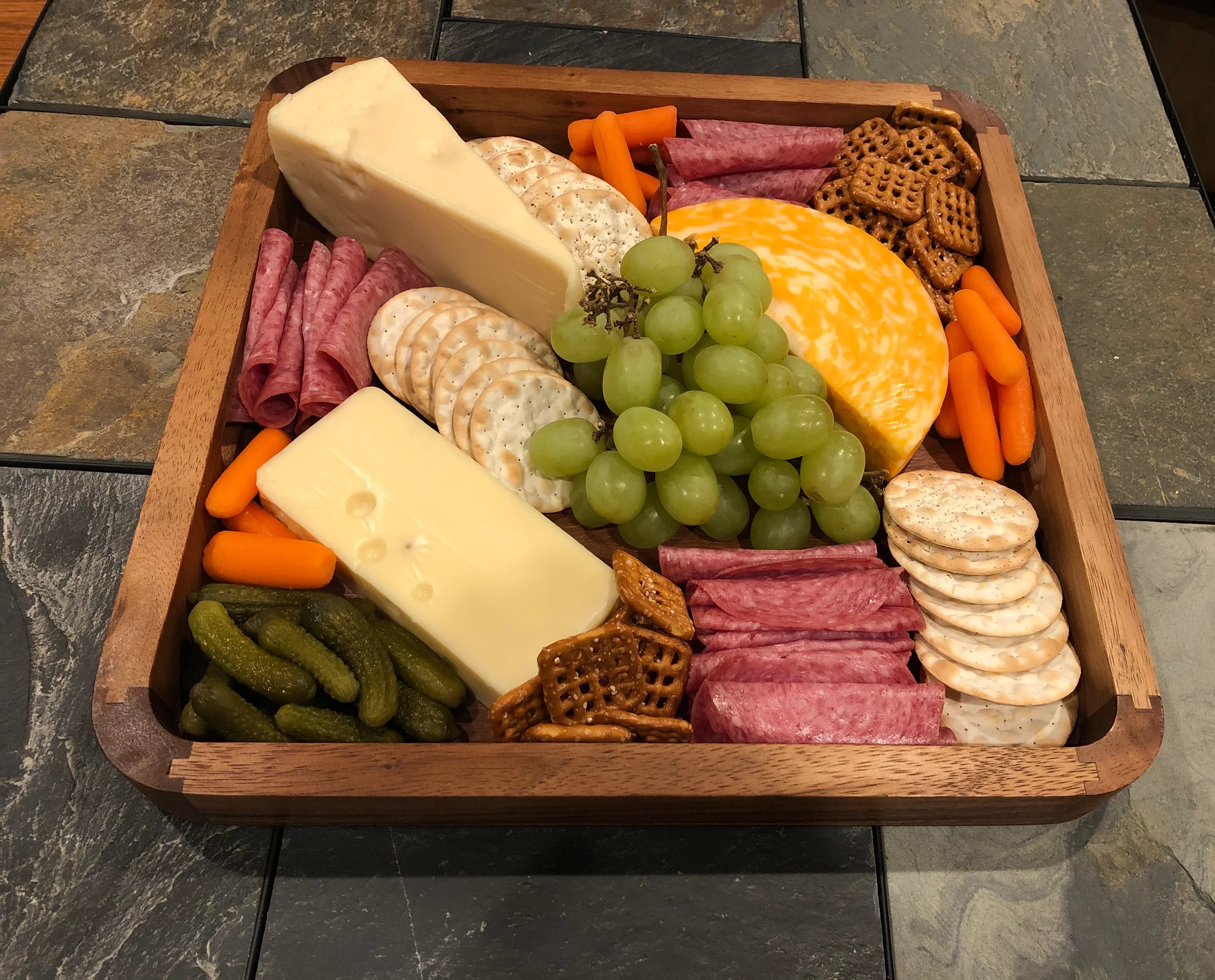 Charcuterie board with assorted cheeses, crackers, salami, green grapes, carrots, pickles, and pretzels.
