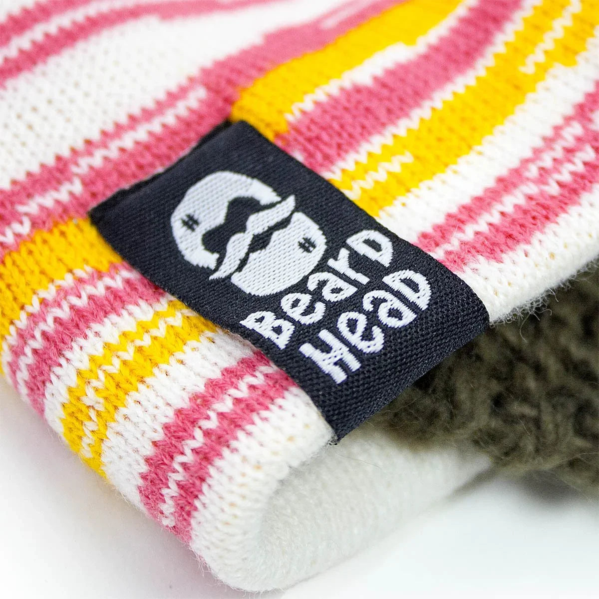 Close-up of colorful striped knit fabric with black Beard Head brand label featuring a white mustached face logo